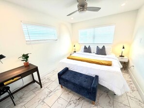 Ein Schlafzimmer mit einem Bett, Nachttischen, einem Schreibtisch, einem Sessel, einem Deckenventilator und einer Zimmerpflanze.