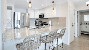 A modern kitchen with stainless steel appliances, a marble countertop, and wicker bar stools. - Oceanfront Two-Bedroom With Large Lanai | Sunset Views, Pool + BBQ (Lahaina)