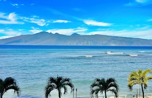 A coastal view with a mountain range in the background, palm trees in the foreground, and a clear blue sky. - Oceanfront Two-Bedroom With Large Lanai | Sunset Views, Pool + BBQ (Lahaina)