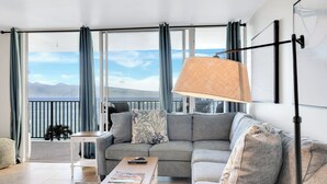 A modern living room with a grey sofa, a coffee table, and a view of the sea through large windows. - Oceanfront Two-Bedroom With Large Lanai | Sunset Views, Pool + BBQ (Lahaina)