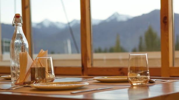 Una mesa de madera para comer con una botella, vasos y cubiertos. Se ve una vista de la montaña a través de amplias ventanas.