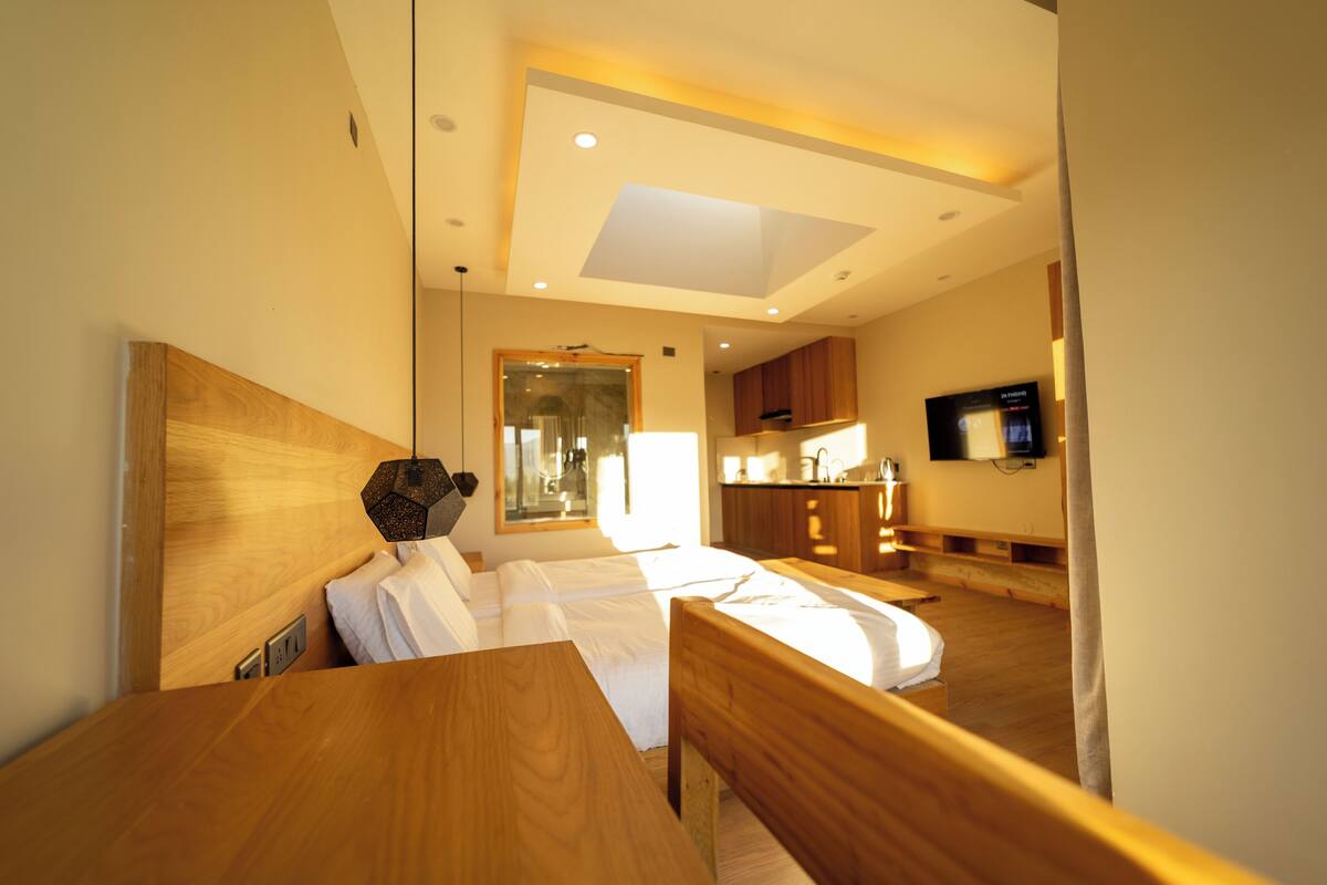 A hotel room with a wooden bed, a small wooden table, a TV mounted on the wall, and a kitchen area visible through an open door.