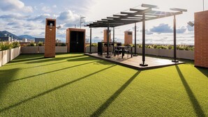 Eine Dachterrasse mit Kunstgrasteppich, einer Pergola, Sitzgelegenheiten im Freien und Blick auf die Stadtlandschaft.