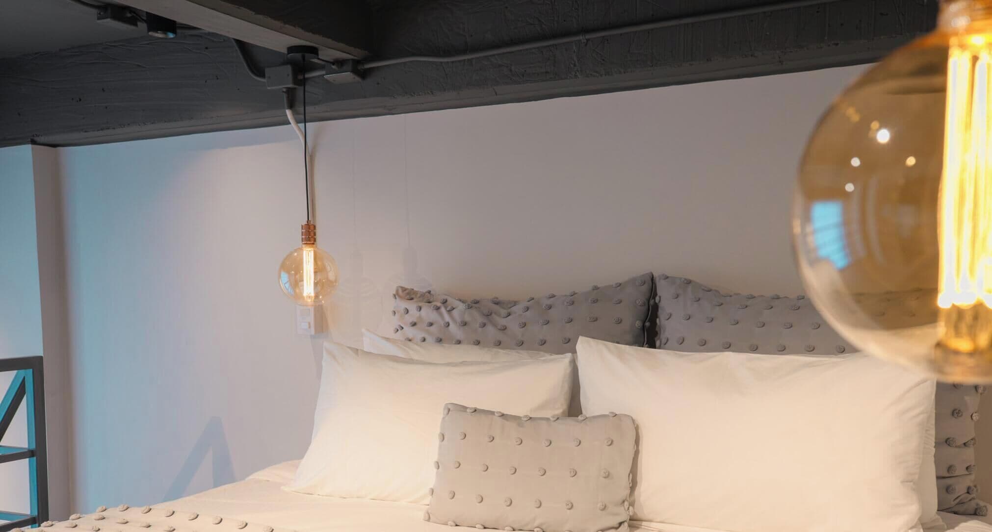 Un letto con biancheria bianca e testiera trapuntata, una lampada a sospensione e un interruttore fissato al muro.