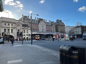 Eine belebte Stadtstraße mit historischen Gebäuden, einer Bushaltestelle und einer Straßenbahnhaltestelle.
