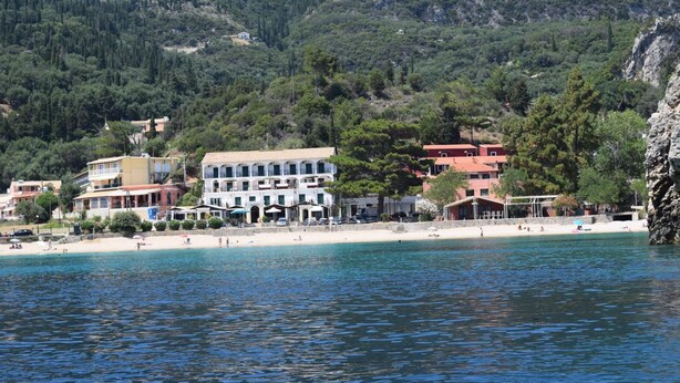 Une ville côtière avec des bâtiments qui bordent la plage, une eau d’un bleu limpide et un paysage montagneux en arrière-plan.
