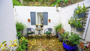En lille terrasse med hvid væg, to stole, et bord og flere krukker med planter.
