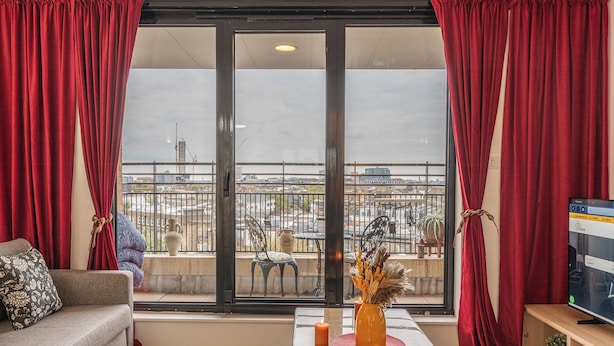 Ein Wohnzimmer mit einer Couch, einem Couchtisch mit Kerzen und einem Fernseher. Es gibt einen Balkon mit Blick auf die Stadt.