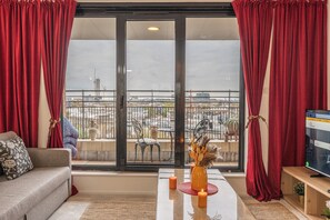 A living room with a sofa, a coffee table with candles, and a TV. There is a balcony with a view of the city. - Duplex 2BR 2BA Flat Near Gloucester Rd Station (London)