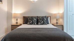 Ein ordentlich gemachtes Bett mit einem gemusterten Bettbezug, zwei passende Kissen und jeweils eine Nachttischlampe auf Holznachtischen.
