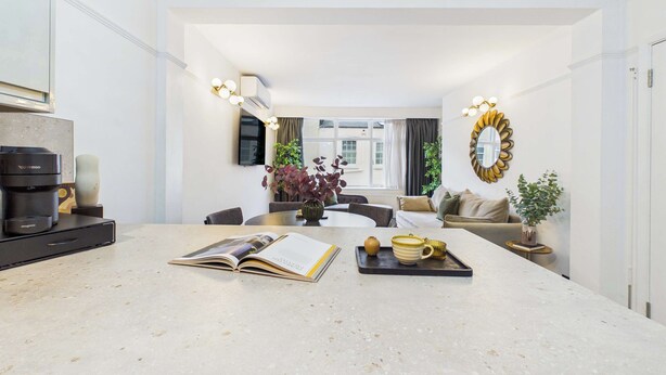 A modern kitchen with a central island, dining area with chairs, and a living space with a sofa and plants. - Elegant Marylebone Mews Apartment (Greater London)