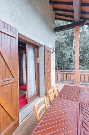 A wooden deck with a table and chairs, an open door leading to a room with a red carpet, and a wooden railing. - Caza Audelou *T5 * Panoramic view of the lake and valley * Fireplace * Parking * (Réal)