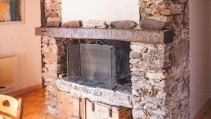 Ein rustikaler Kamin aus Stein mit einem Metallgitter, Holzbalken und einem Kachelboden.