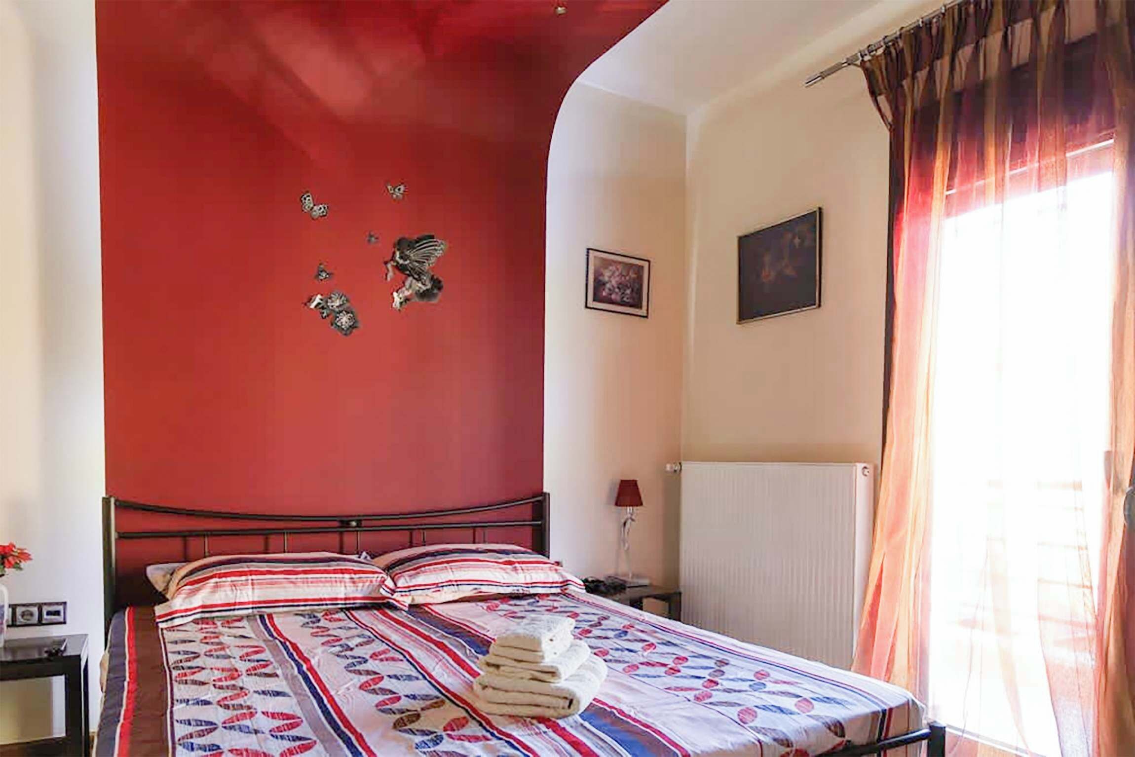 Un dormitori amb una paret d'accent vermella, un llit amb llençols estampats, un radiador i una finestra amb cortines.