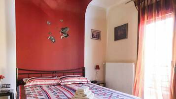 Un dormitori amb una paret d'accent vermella, un llit amb llençols estampats, un radiador i una finestra amb cortines.