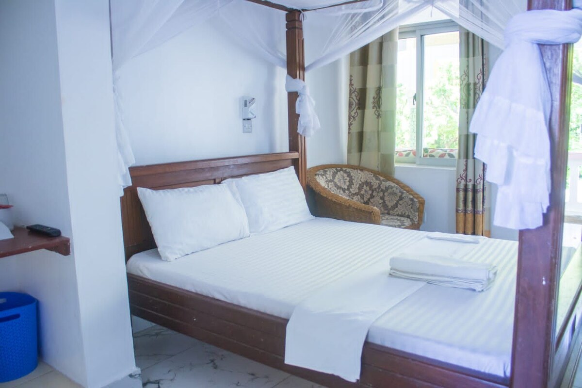 A bed with a canopy, a bedside table, a chair, and a window with curtains.