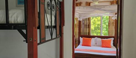A room with a bunk bed, a single bed with orange pillows, and a view of the outside through a doorway.