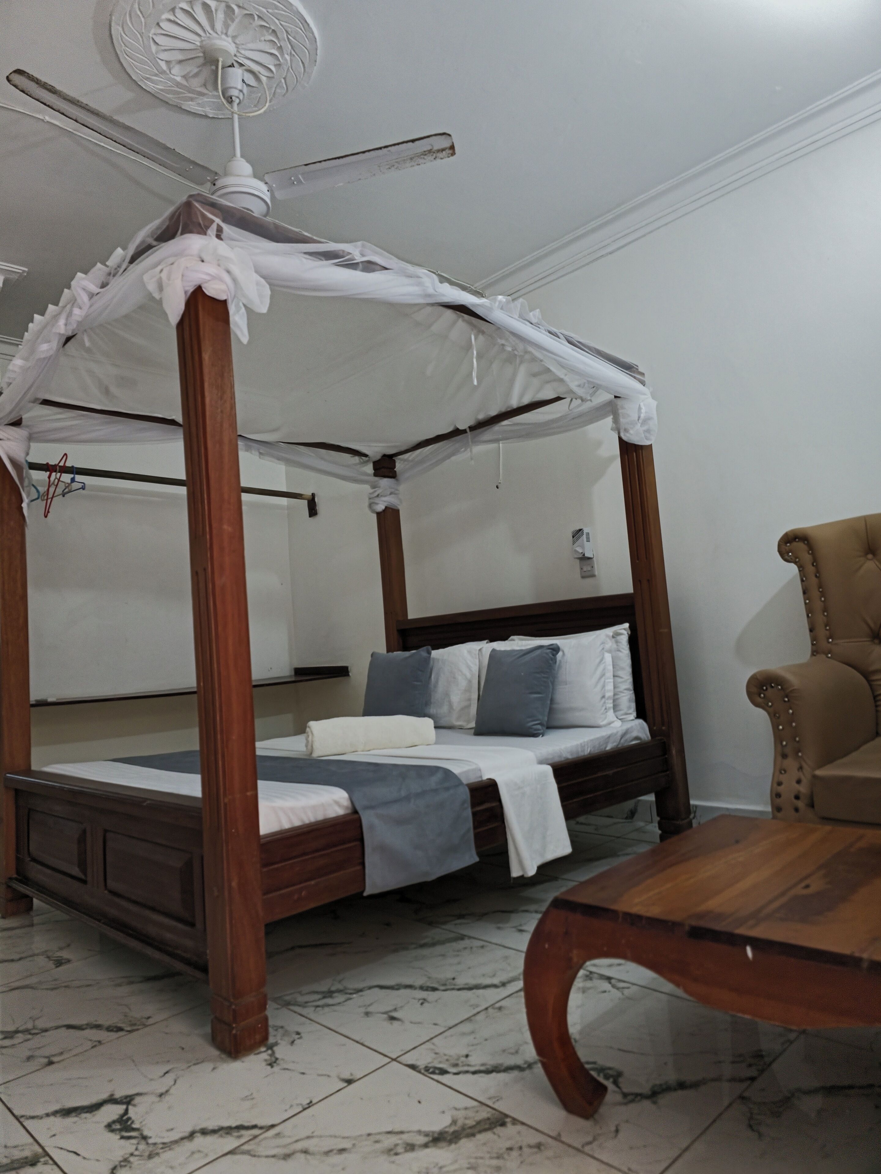 A four-poster canopy bed with a wooden frame and white bedding, a wooden chair, and a marble-tiled floor.
