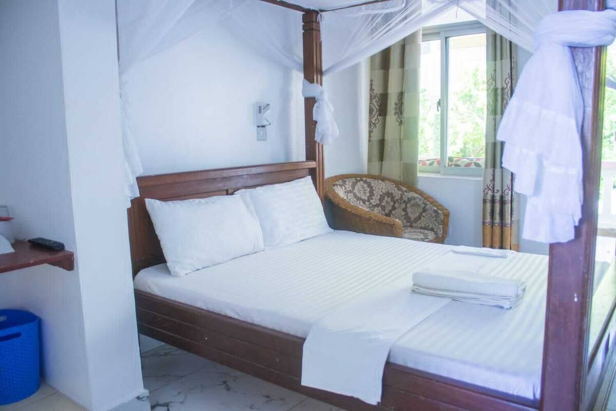 A bed with a canopy, a bedside table, a chair, and a window with curtains.