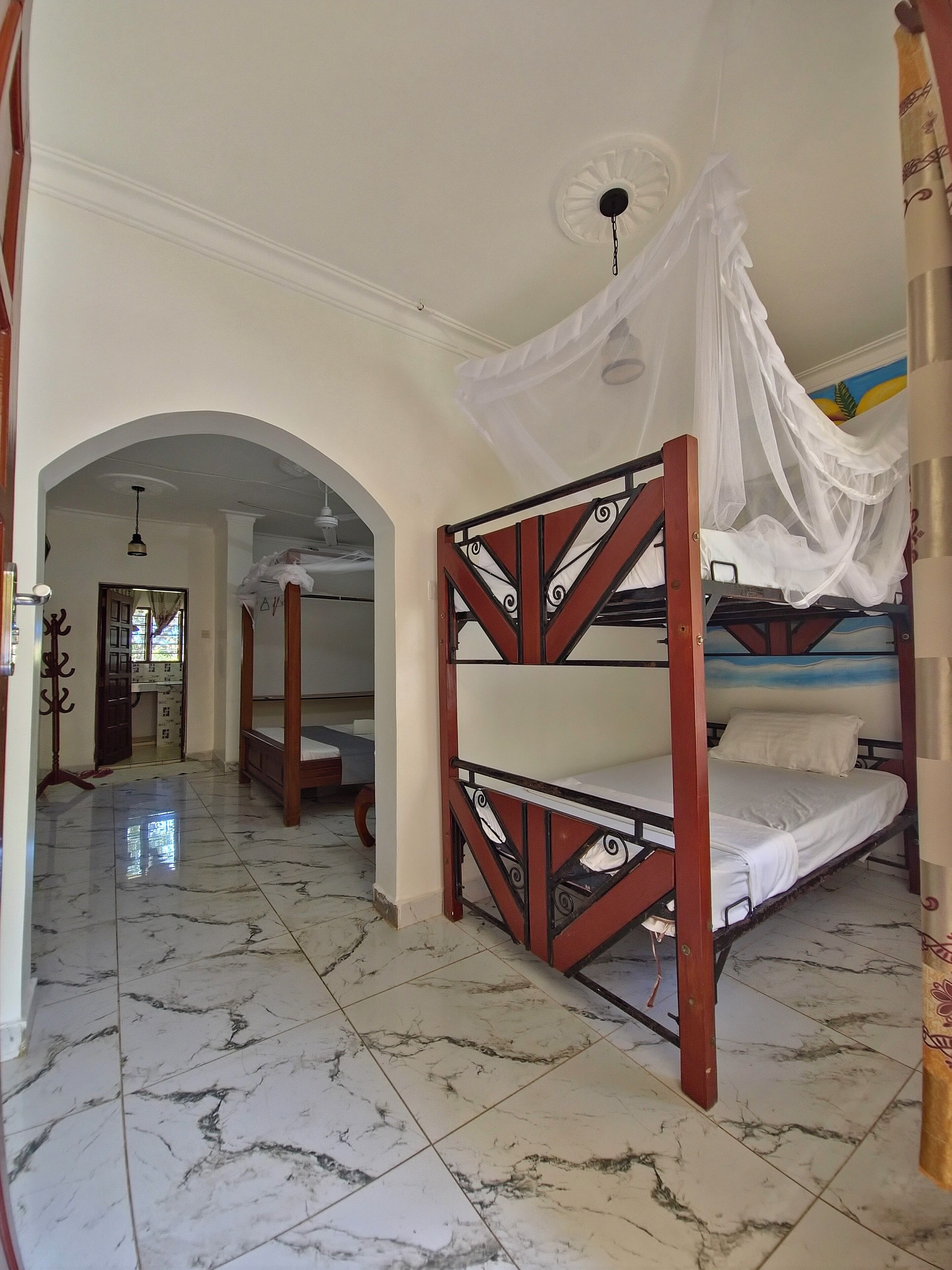 A room with a bed under a canopy, a marble-tiled floor, and a hallway with an arched doorway leading to another room.