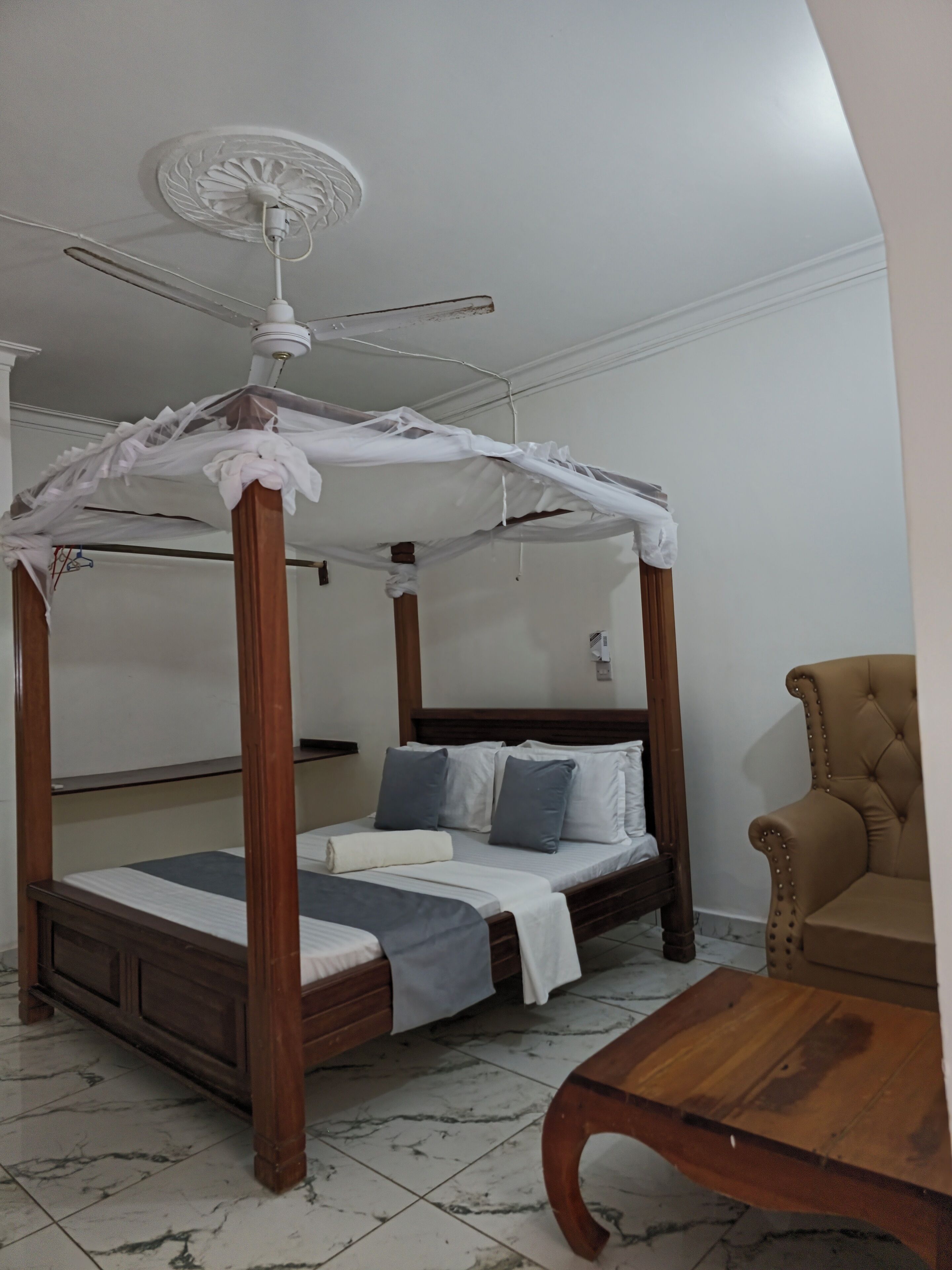Uma cama com dossel e cortina, um ventilador de teto, uma mesa de cabeceira de madeira e uma poltrona estofada.