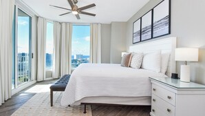 A modern bedroom with a large bed, a ceiling fan, a nightstand, and a view of the city through floor-to-ceiling windows. - The Palms of Destin 21117 by Compass Resorts (Destin)