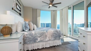 A bedroom with a large bed, a ceiling fan, a nightstand with a lamp, and a view of the city through large windows. - The Palms of Destin 21117 by Compass Resorts (Destin)