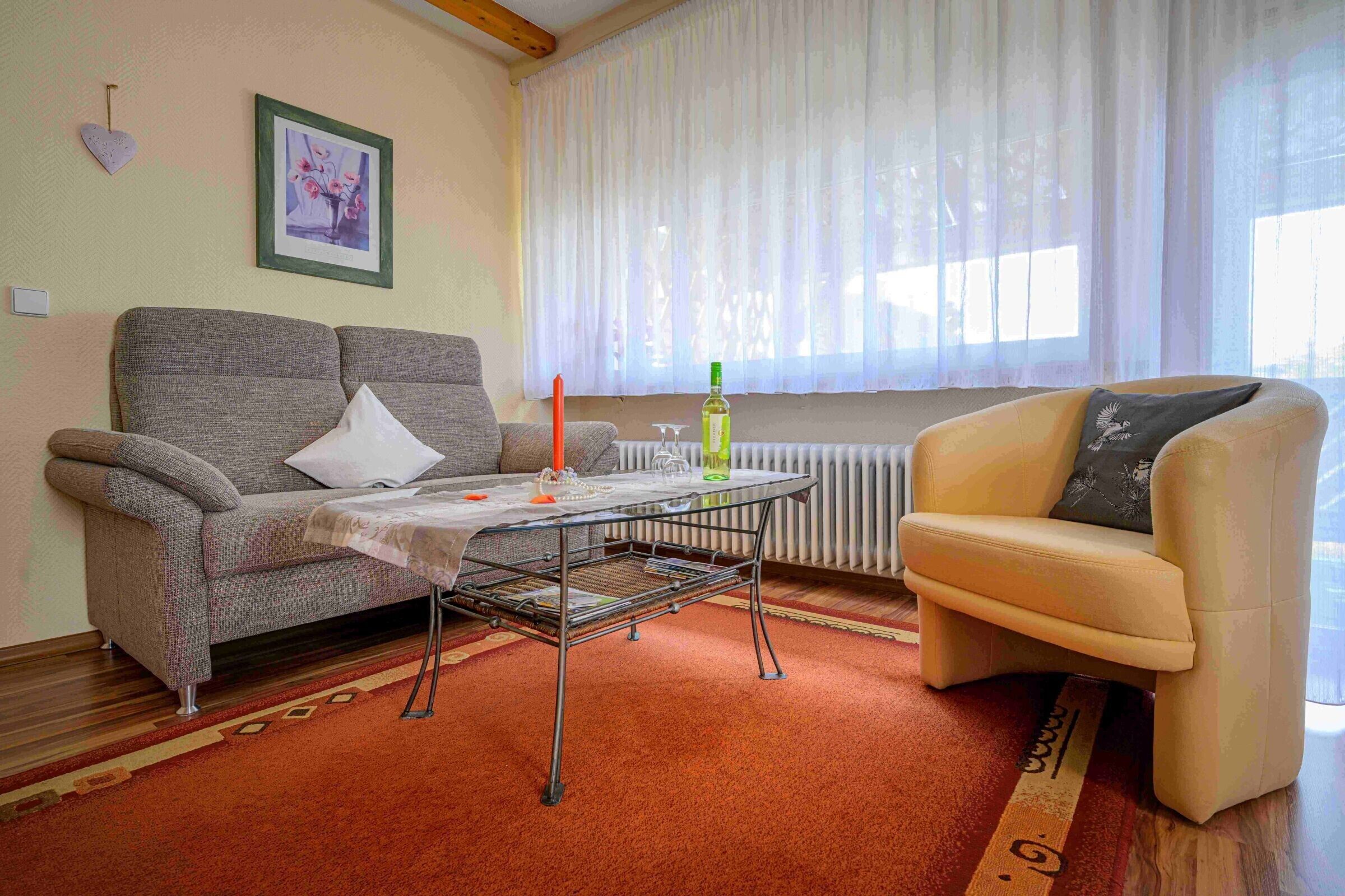 Ein Wohnzimmer mit einer Couch, einem kleinen Tisch, einem Sessel, einer Heizung und einem Fenster mit leichtem Vorhang.