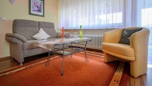 A living room with a sofa, a small table, a chair, a radiator, and a window with sheer curtains. - Apartment 'Ferienwohnung Doll Waltraud' with Shared Terrace and Balcony (Oberkirch)