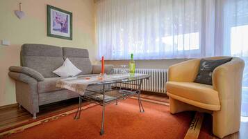 Ein Wohnzimmer mit einer Couch, einem kleinen Tisch, einem Sessel, einer Heizung und einem Fenster mit leichtem Vorhang.