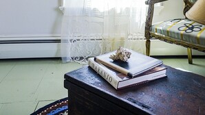 A room with a large window covered by sheer curtains, a framed picture on the wall, a chair with a floral cushion, and a table with books and a seashell.