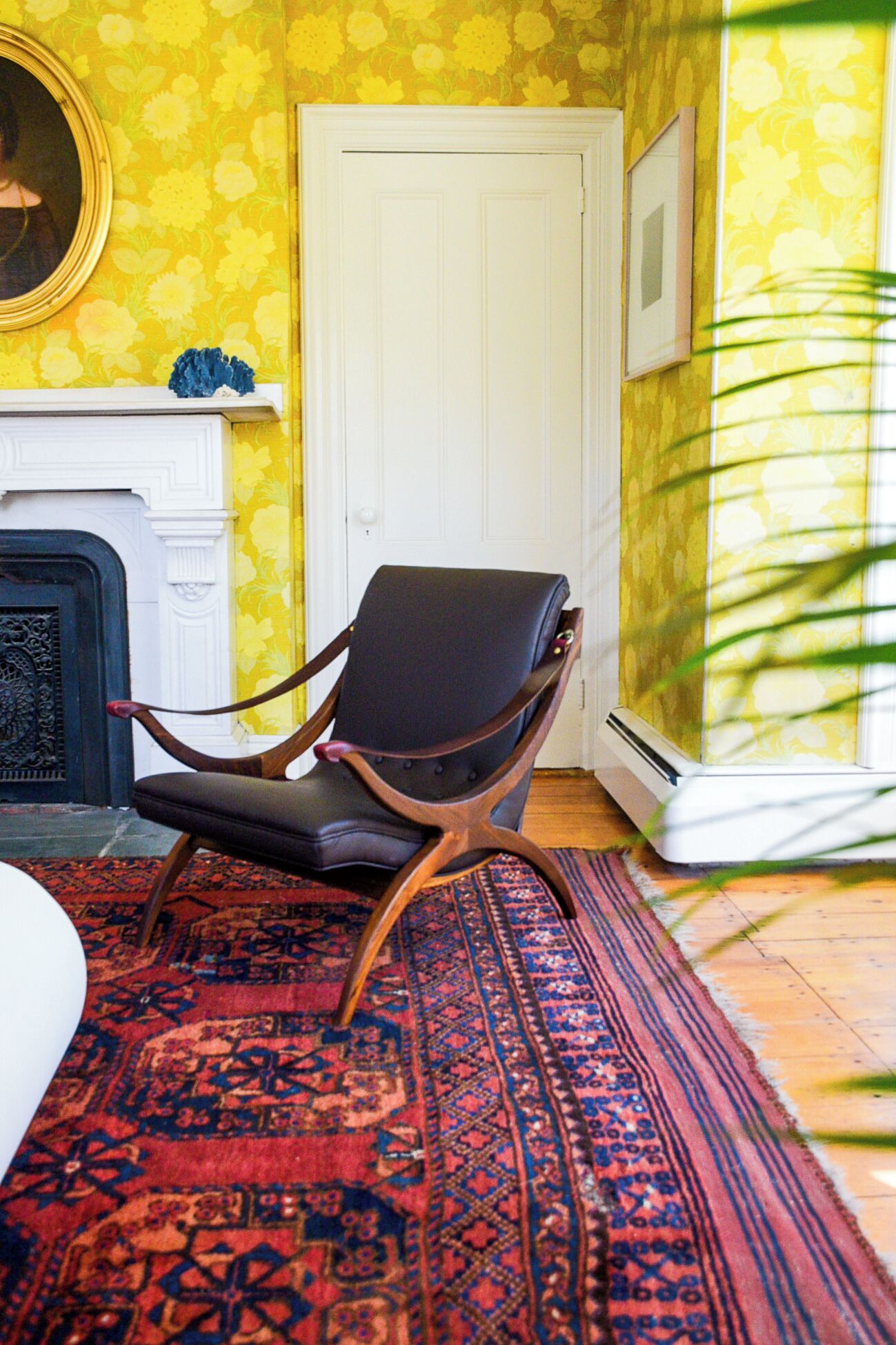 A room with a patterned yellow wall, a white door, a black leather chair, a red and blue patterned rug, and a white fireplace.