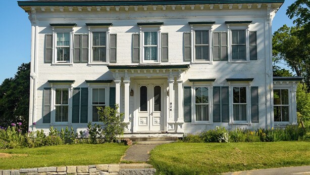 A two-story white building with green shutters, a central entrance with columns, and a brick chimney.