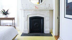 A bedroom with a fireplace, a round mirror, a bed, a bedside table, and a window with curtains.