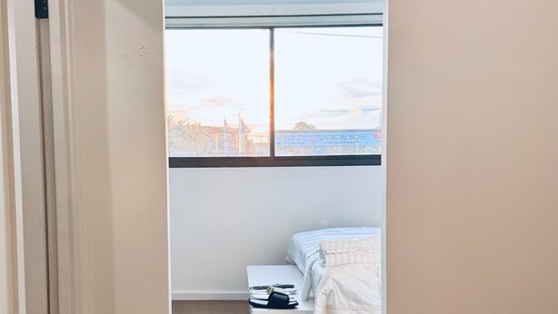 Ein schmales Zimmer mit einem Bett, einem kleinen Tisch und einem Fenster mit Blick auf Gebäude.