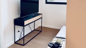 Ein modernes Zimmer mit einem schwarzen Fernseher auf einem schlichten Standfuß, einer Wanduhr und einem Fenster mit Blick ins Freie.