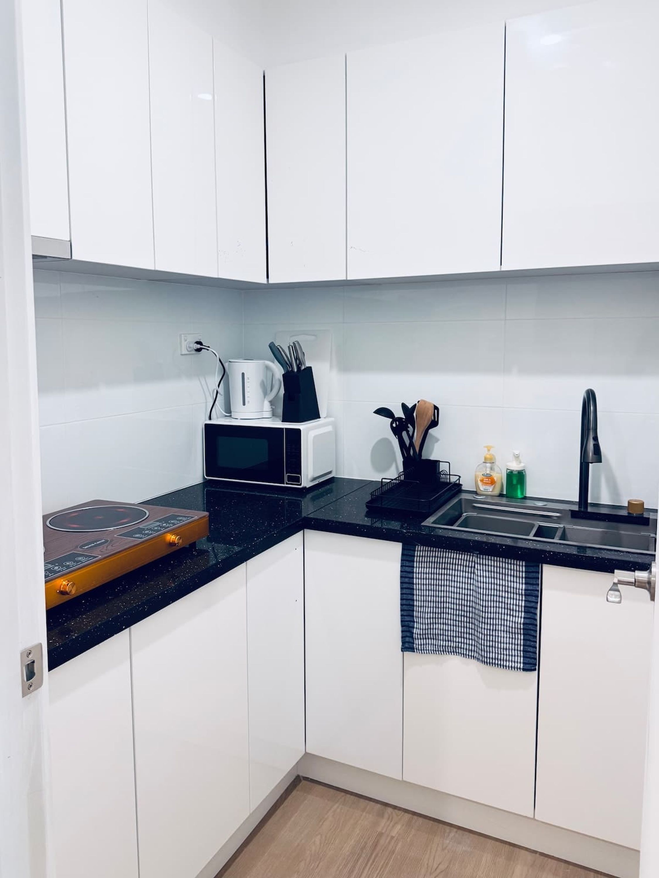 Una cocina pequeña con armarios blancos, encimera negra, microondas, hervidor de agua, cafetera, fregadero y cocina de inducción de madera.