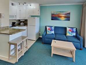 A compact living space with a kitchenette, a blue sofa with colorful cushions, and a painting of a sunset. - Casa Del Mar 387 | Partial Gulf Views, Pool Access (Galveston)