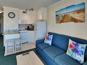 A compact living space with a kitchenette, a blue sofa with colorful cushions, and a beach-themed painting on the wall. - Casa Del Mar 323 | Partial Gulf Views (Galveston)