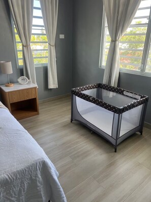 Ein Schlafzimmer mit Bett, Nachttisch, einem Babyreisebett mit Glasplatte und einem Fenster mit Vorhängen.