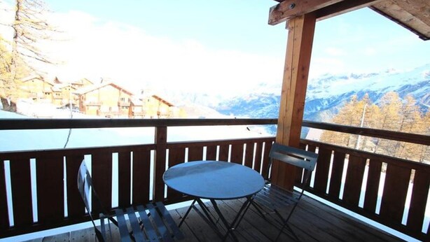 Une terrasse en bois avec une table et des chaises, offrant une vue sur un paysage montagneux parsemé de bâtiments.