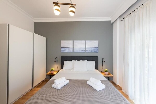 A modern hotel room with a large bed, a grey wall, a view of the sea, and a ceiling light fixture. - PSV Mitropoleos (Thessaloniki)