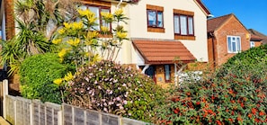 A residential building with a red-tiled roof, multiple windows, and a variety of plants and shrubs in the surrounding garden.