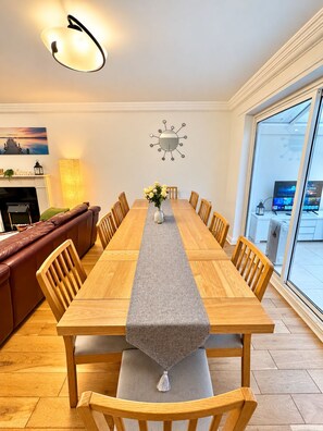 Uma sala de jantar com uma mesa de madeira comprida, cadeiras acolchoadas, um vaso com flores, um relógio de parede e uma lareira.