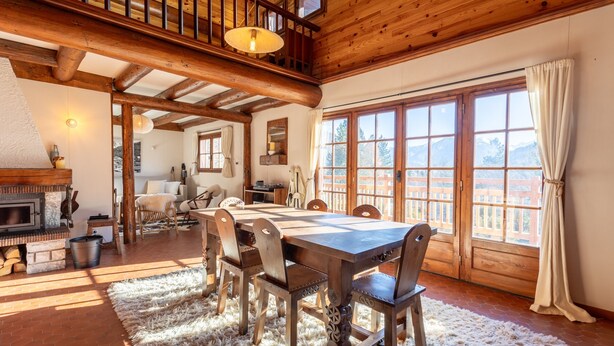 A spacious dining area with a wooden table and chairs, a fireplace, and large windows with curtains.