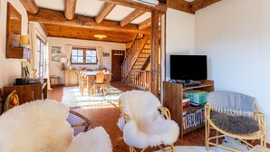 A rustic interior with exposed wooden beams, a dining area, a staircase, and a television.