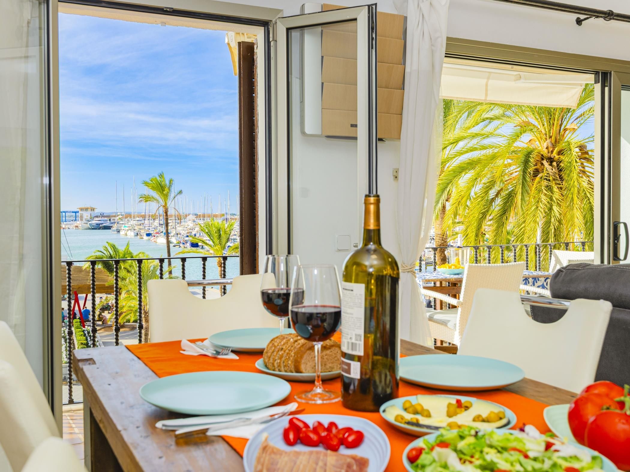 A dining table with plates, glasses, and a bottle, overlooking a marina with boats and palm trees.