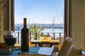 Un comedor con una mesa dispuesta para dos personas, una botella de vino y una copa de vino tinto. La mesa está ubicada cerca de una ventana con vistas a un puerto deportivo lleno de barcos.