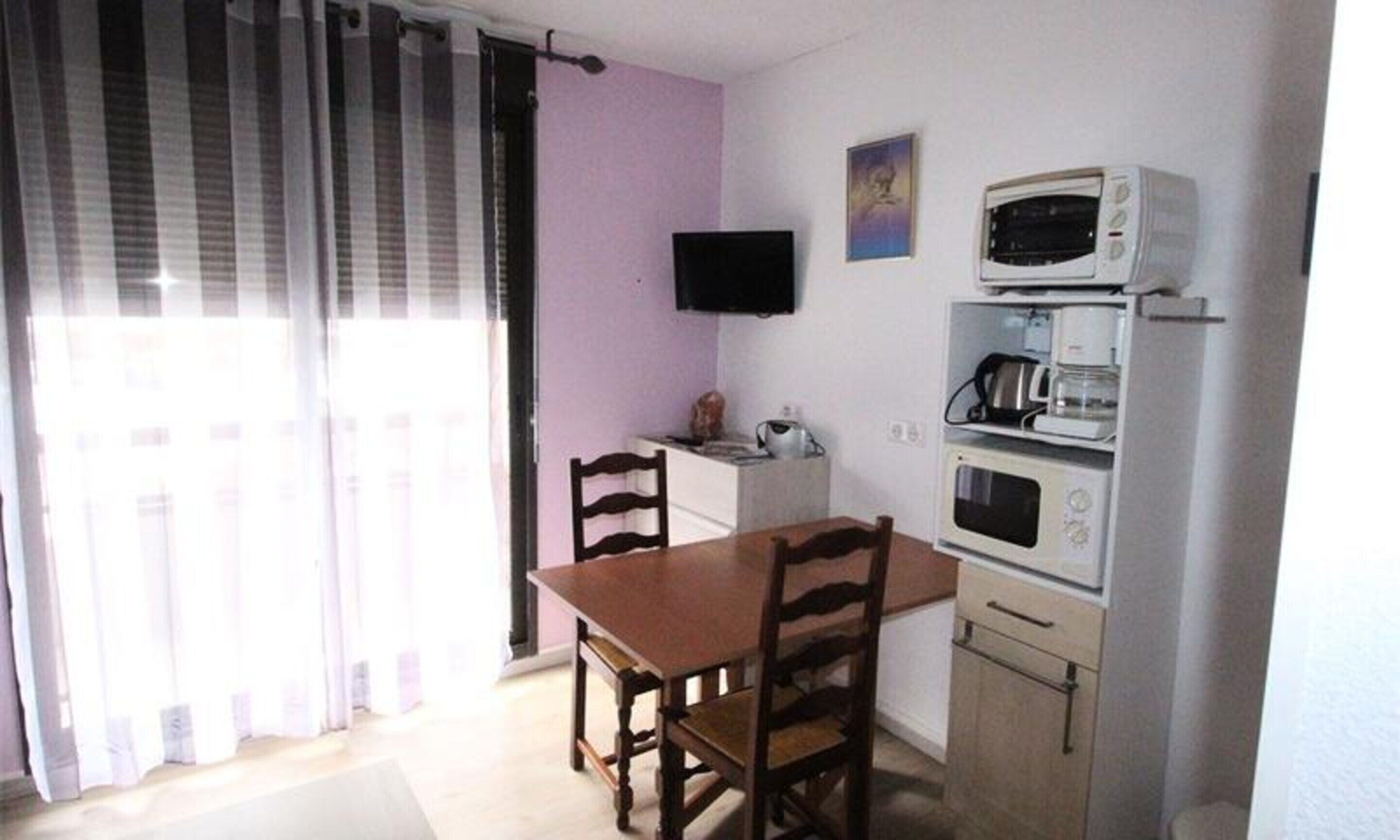 Een lichte, compacte kamer met een houten eettafel en stoelen, een flatscreen televisie aan de muur en een kitchenette met een magnetron en koffiezetapparaat.