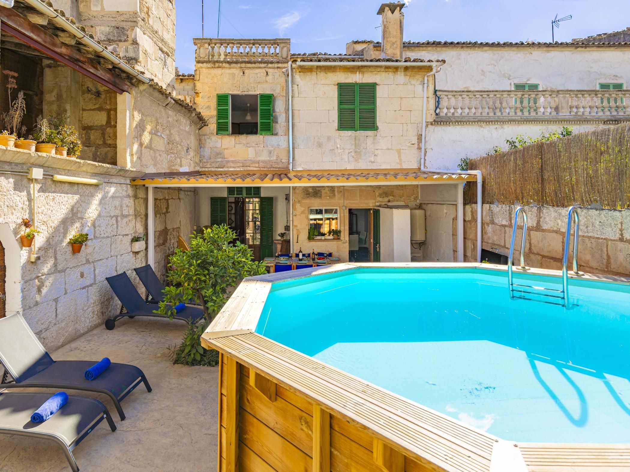 A pool area with a wooden deck, lounge chairs, and a stone building with green shutters.
